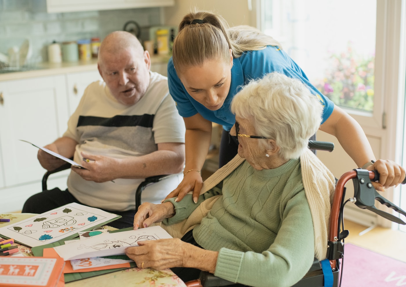 A caregiver sitting with two older adults at home, looking at something together in warm light